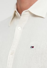 Λευκό πουκάμισο από λινό με κουμπιά, με μυτερό γιακά, κουμπιά μπροστά και μικρό κεντητό λογότυπο σε κόκκινο, άσπρο και μπλε στο στήθος.