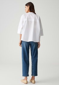 Witte oversized blouse met 3/4 mouwen, knoopmanchetten en een rugpaneel detail; gecombineerd met hoge jeans in blauw en beige platte schoenen.