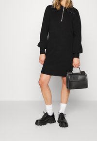 Svart stickad klänning med dragkedja i kragen, texturerade ärmar, kombinerad med kraftiga svarta lackskor, vita ankelsockor och en strukturerad svart handväska.