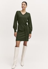 Abito a maglia verde con maniche lunghe e scollatura a V, dotato di una cintura annodata in vita. Abbinato a stivali bianchi al ginocchio con punta.
