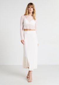 Donna che indossa un crop top lungo a maniche in pizzo bianco, un bralette bianco, una lunga gonna bianca e sandali con tacchi alti bianchi, in piedi su uno sfondo neutro.