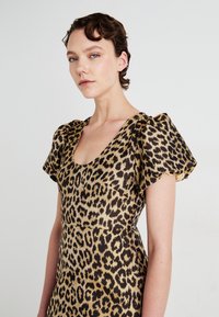 Žena s krátkymi hnedými vlasmi, oblečená v šatách s balónovými rukávmi a leopardím vzorom, ktorá sa jemne otáča doľava na svetlom pozadí.