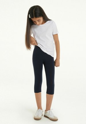 Maglietta bianca a maniche corte, leggings capri blu scuro e scarpe bianche con dettagli beige; modella che guarda in basso, capelli parzialmente raccolti con una clip.