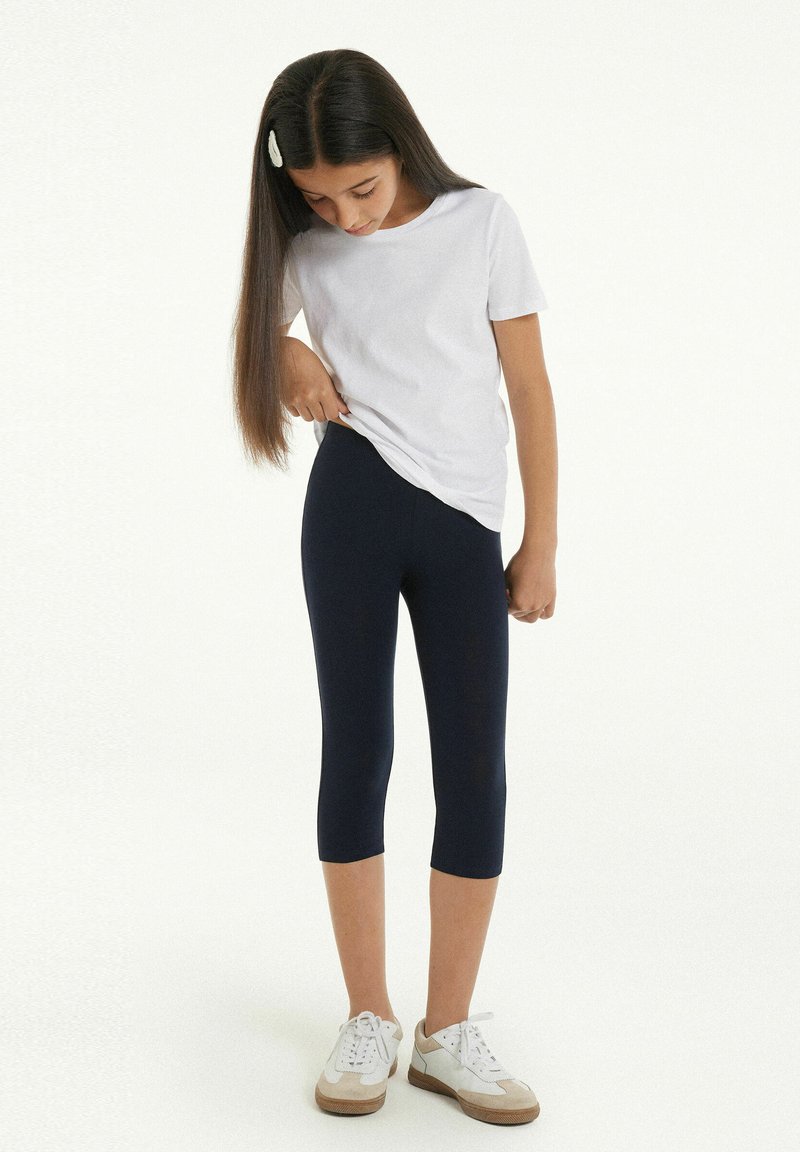 Bílý tričko s krátkým rukávem, námornickomodré legíny do pasu a bílé boty s béžovými akcenty; modelka se dívá dolů, vlasy částečně drženy sponou.