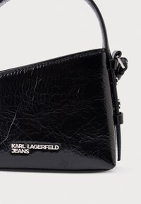 Melna, krunkaina ādas rokassomiņa ar īsu rokturi un sudrabotu logotipu "KARL LAGERFELD JEANS" priekšpusē. Vienkāršs, leņķisks dizains.