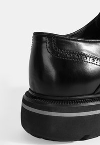 Zwarte leren schoen met een brogue-detailing bij de hak, met een dikke zwarte zool en een contrasterende grijze streep langs de onderkant.