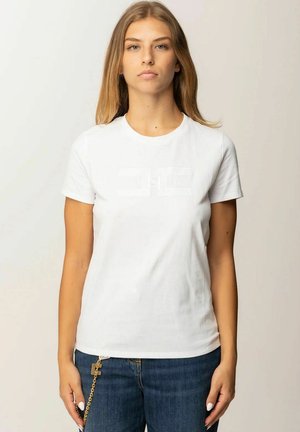 Giovane donna con lunghi capelli biondi indossa una maglietta bianca semplice a maniche corte e jeans blu con un accessorio a catena dorata.