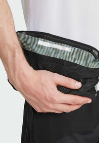 Riñonera de malla negra con una sección superior de patrón geométrico, que cuenta con una cremallera y una etiqueta blanca con la marca "TERREX", sostenida en una mano.