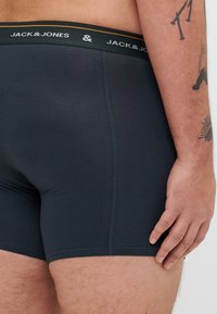 Dunkelgraue Boxershorts mit elastischem Bund, der das Logo "JACK & JONES" in Weiß und Orange zeigt. Glattes Gewebe, enganliegendes Design, kurze Beine.