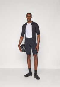 Schwarzes Radtrikot mit kurzen Ärmeln, weißes Mesh-Unterhemd, schwarze gepolsterte Shorts und Radschuhe; hält einen schwarzen Helm.