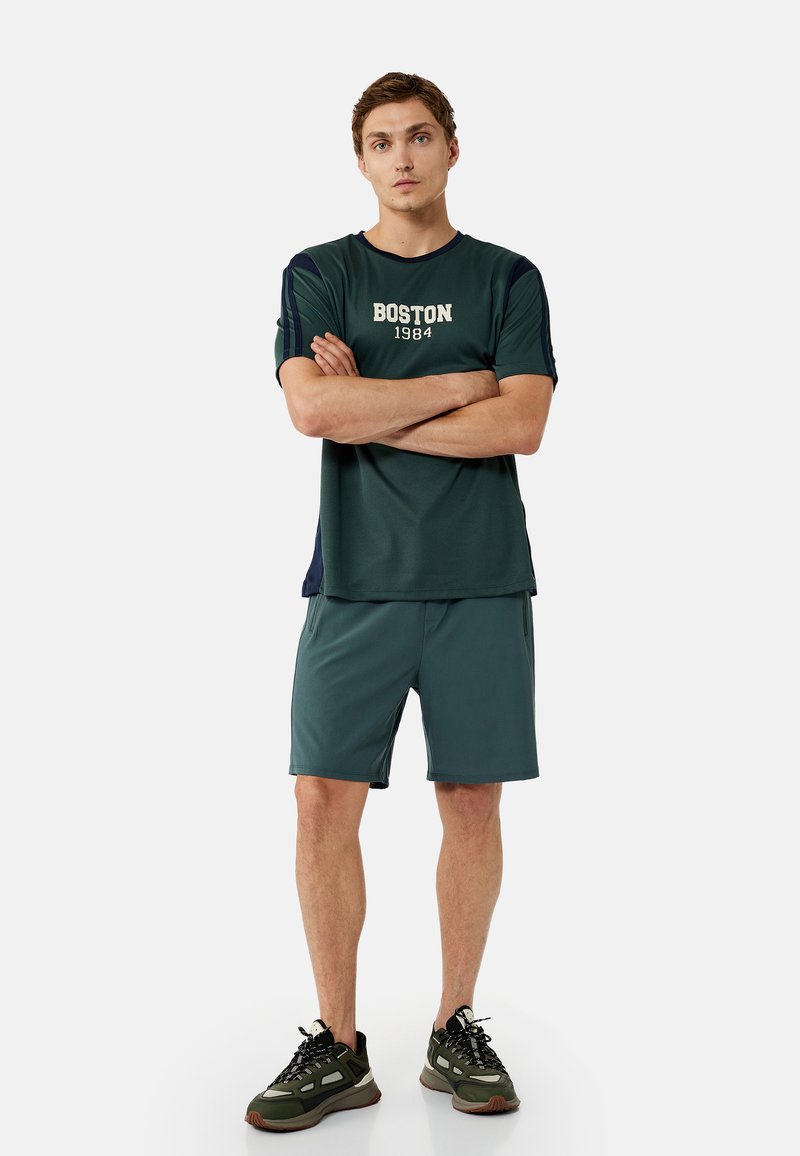Young man standing with arms crossed wearing dark green "Boston 1984" t-shirt, matching shorts, and green sneakers on white background.