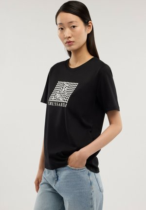 Joven con cabello lacio y negro que lleva una camiseta negra con un diseño gráfico y jeans azul claro, posando con una mano en el bolsillo.