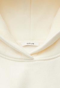 Gros plan sur l'encolure d'un sweat à capuche écru montrant l'étiquette de marque cousue "OPUS" à l'intérieur du col.