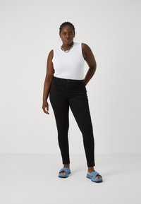 Svarta slim jeans i kombination med en åtsittande vit linne och blå sandaler. Jeansen har fickor och knappstängning.