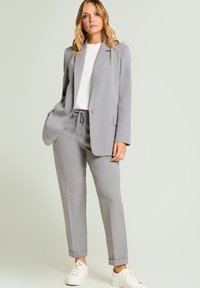 Grauer, maßgeschneiderter Blazer mit feinem Punktmuster, kombiniert mit passenden, schmal zulaufenden Hosen und weißen Sneakern. Glatter Stoff mit einer strukturierten Gestaltung.