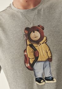 Szürke pulóver, amelyen egy sárga kapucnis pulcsis medve grafikája látható, aki gitározik, farmernadrág és fekete sportcipő kék részletekkel.