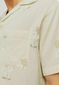 Beige Kurzarmhemd mit strukturiertem Muster, einer Brusttasche und floralen Drucken in gedämpften Farben.