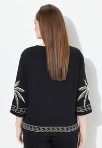 Top negro con mangas 3/4 y bordado de palmera beige, además de un patrón decorativo de olas en el dobladillo y los puños de las mangas. Textura de tela suave.