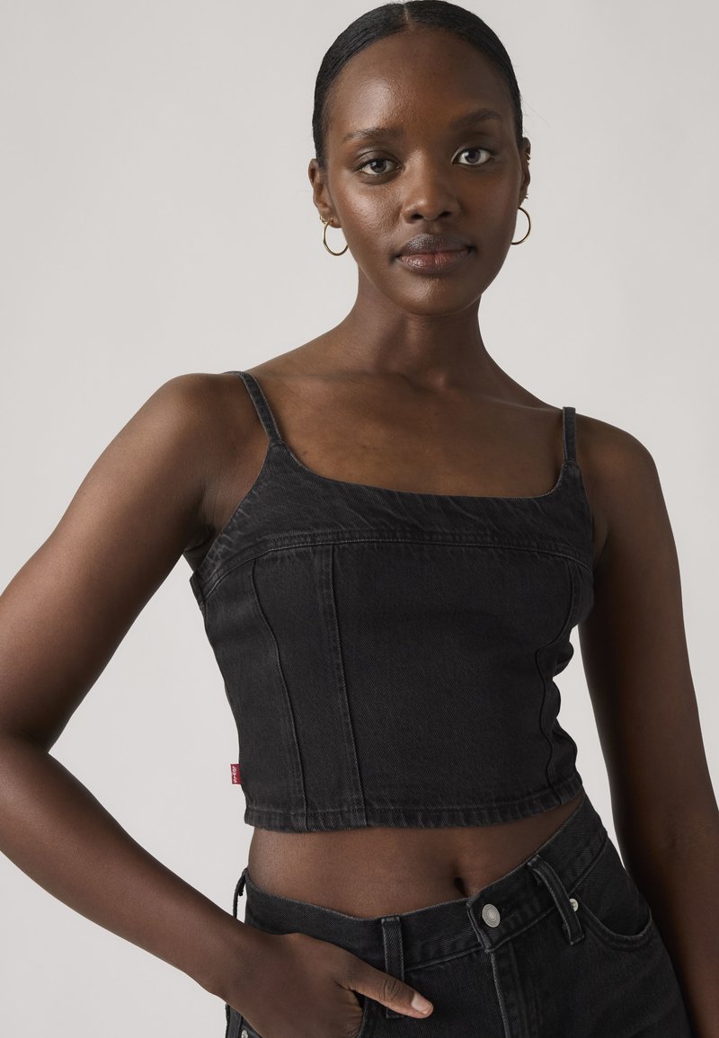 Jeune femme à la peau foncée portant un crop top en jean noir et un jean taille haute assorti, debout devant un fond clair uni.