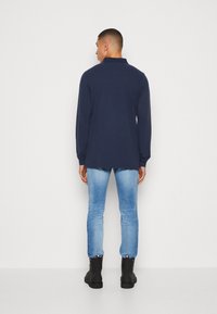 Polo shirt a maniche lunghe blu navy con colletto, abbinato a jeans skinny blu chiaro e stivaletti neri alla caviglia, visto da dietro.