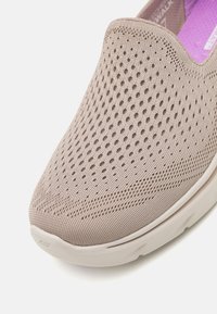 Sandalho bege com um cabedal de malha que apresenta buracos de ventilação circulares, uma superfície texturizada e uma sola almofadada de cor clara.