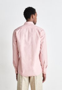 Camicia button-up rosa chiaro con maniche lunghe, orlo arrotondato e texture sottile. Presenta dettagli con polsini abbottonati per la chiusura.