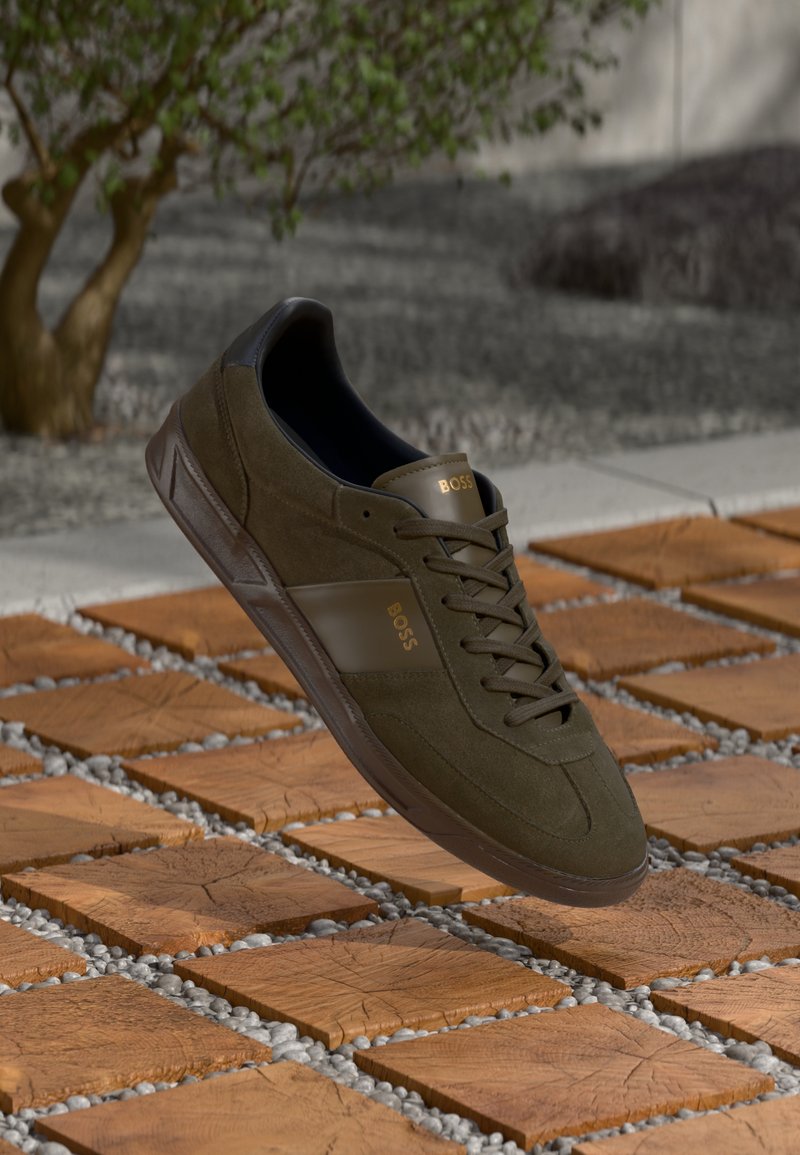 Mørkegrønne semskede sneakers med sorte detaljer og gullogo, lavt design, snøring, lett s�åle, vist på teksturert flisoverflate.