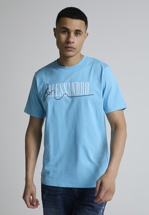 Ung mann iført en lys blå T-skjorte med teksten "ALESSANDRO" og svart design, stående foran en enkel grå bakgrunn.