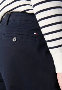 Mörkblå byxor i bomull med en bakficka som har en märkesknapp och en liten tric färgad logotypaccent. Rutig tröja synlig.