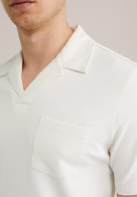 Tricou polo de culoare crem, cu mâneci scurte, guler tip șal, un buzunar pe partea stângă a pieptului și textură netedă.