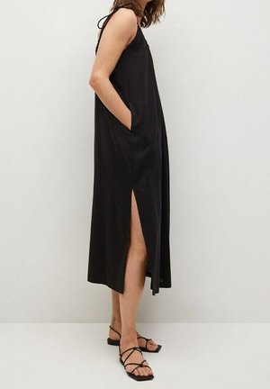 Frau trägt ein ärmelloses schwarzes Midikleid mit seitlichen Taschen und einem hohen Schlitz, kombiniert mit schwarzen flachen Sandalen mit Riemchen.