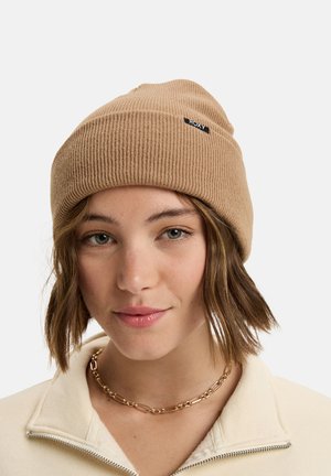 Tan ribbed knit beanie with a small black logo. Worn with a cream zip-up fleece and gold chain necklace, featuring a smooth texture.
