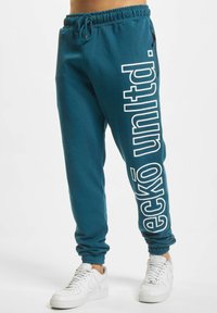 Kalkblå bomullssweatpants med snor i midjen, smale mansjetter og hvit "eckō unlt d." tekst på venstre side. Sett sammen med hvite sneakers.