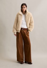 Femme portant une veste en sherpa beige, une chemise blanche à zip, un pantalon large marron, une ceinture marron et des bottes à talons marron, se tenant devant un fond uni.