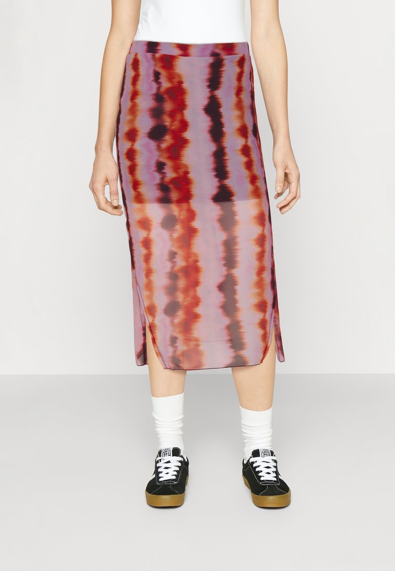 Femme portant une jupe midi tie-dye violet, rouge et noir avec des fentes sur les côtés, des chaussettes blanches et des baskets noires avec des semelles en caoutchouc.