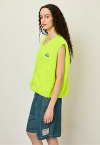 Bright lime green fleece vest with a zip closure, sleeveless design, front zip pockets, and a small blue brand patch. Paired with denim shorts.
