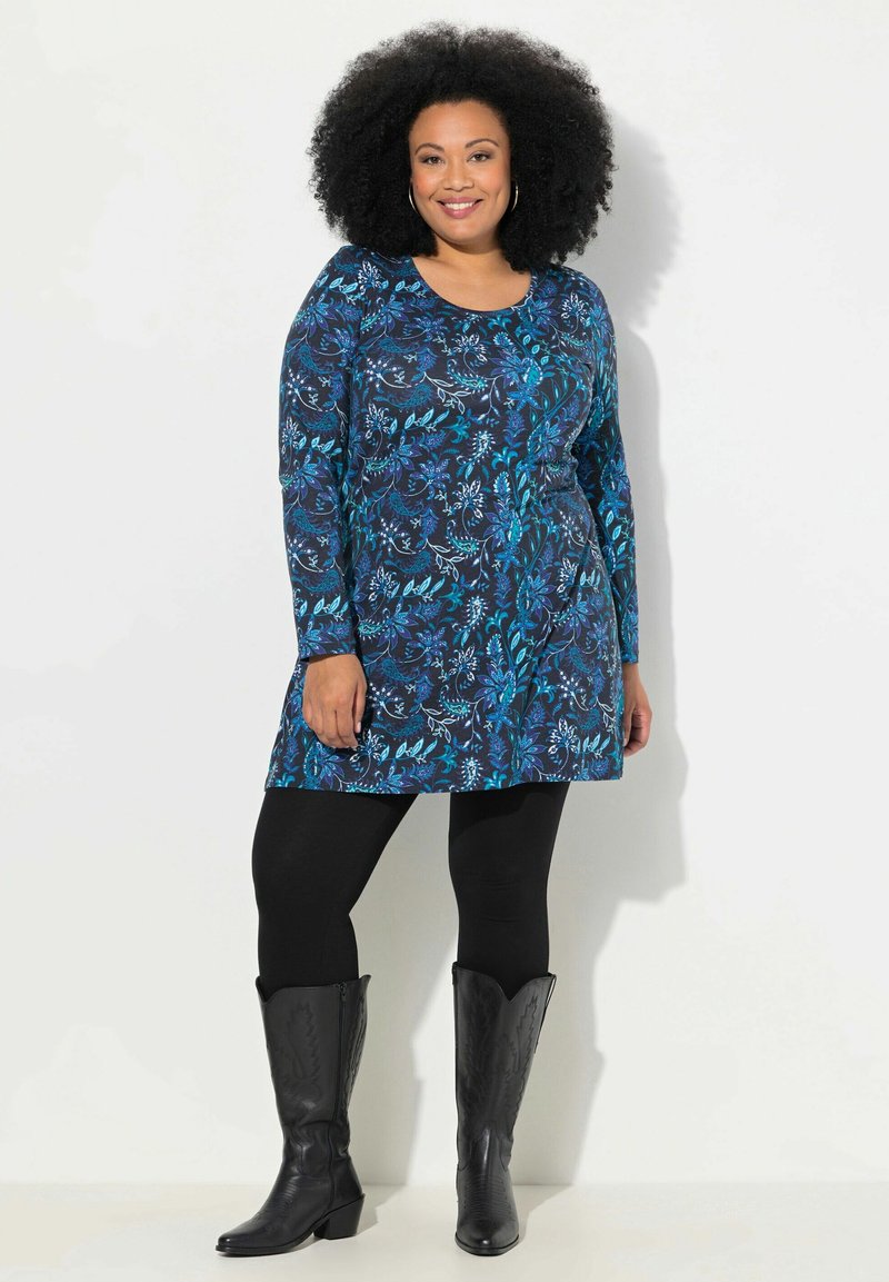 Langsleeve jurk in donkergroen met bloemmotieven in turquoise. Draagt over zwarte leggings en gecombineerd met zwarte cowboylaarzen.