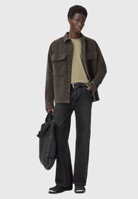 Giacca di velluto a coste marrone con due tasche frontali, t-shirt verde, jeans neri e scarpe slip-on nere. Tieni in mano una borsa grigia con una tracolla.