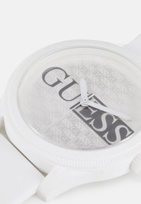 Montre-bracelet en silicone blanc avec un cadran transparent comportant le logo "Guess" en motif répétitif, des aiguilles blanches et une lunette texturée.