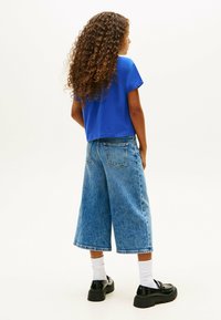 Camiseta azul cropped combinada con pantalones vaqueros de cintura alta de mezclilla azul claros. Botas negras gruesas y calcetines blancos. Pelo rizado visible desde atrás.