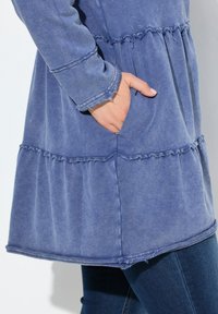 Abito blu in misto cotone leggero con design a volant a strati, maniche lunghe e tasca frontale. Rifinito con bordi grezzi.