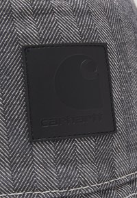 Graues Fischgrätgewebe mit einem strukturierten schwarzen Lederpatch, auf dem ein kreisförmiges Logo und das Wort "carhartt" eingepresst sind.