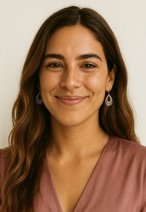 Lächelnde Frau mit langen, welligen braunen Haaren, die ein mauvefarbenes V-Ausschnitt-Top und silberne Tropfenohrringe trägt, vor einem schlichten, cremefarbenen Hintergrund.