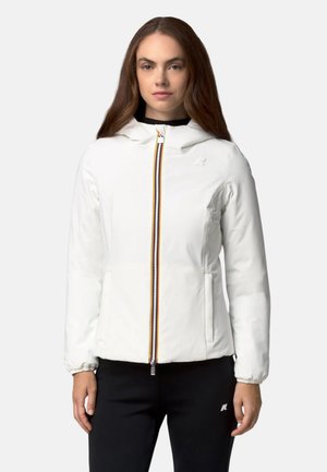 Une femme porte une veste blanche à capuche avec une fermeture éclair multicolore sur le devant et un pantalon noir, sur un fond uni.