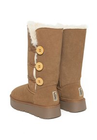 Botas de ante marrón con forro de piel de oveja, que presentan tres botones de madera redondos y una suela de goma texturizada. Detalle del logotipo en la parte trasera.