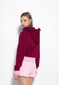 Czerwony hoodie z falbaniastym detalem przy kapturze, w połączeniu z jasnoróżowymi szortami. Ubranie ma kontrastujące prążkowane mankiety i pas. 