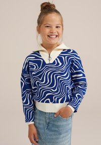 Kék és fehér pulóver, hullámos mintázattal, félig zipzáros gallérral és bordás szegéllyel. Egy világoskék farmer szoknyával párosítva.