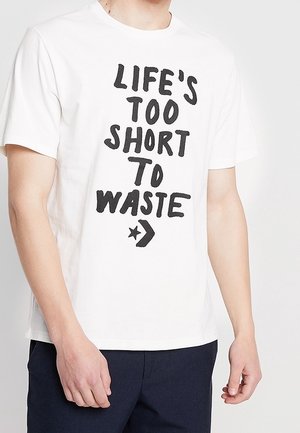 Hombre con una camiseta blanca con texto negro que dice "LA VIDA ES DEMASIADO CORTA PARA DESPERDICIARLA" y un pequeño símbolo de estrella y flecha debajo del texto.