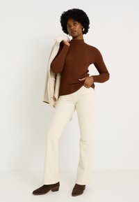 Pull à col roulé marron à côtes avec un motif vertical, pantalon évasé beige clair, et bottines en daim marron. Veste grise drapée sur l'épaule.