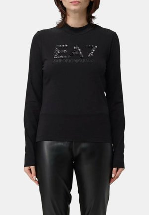 Donna che indossa una maglia nera a maniche lunghe con la scritta "EA7 Emporio Armani" in paillettes e pantaloni di pelle neri, in piedi davanti a uno sfondo uniforme.